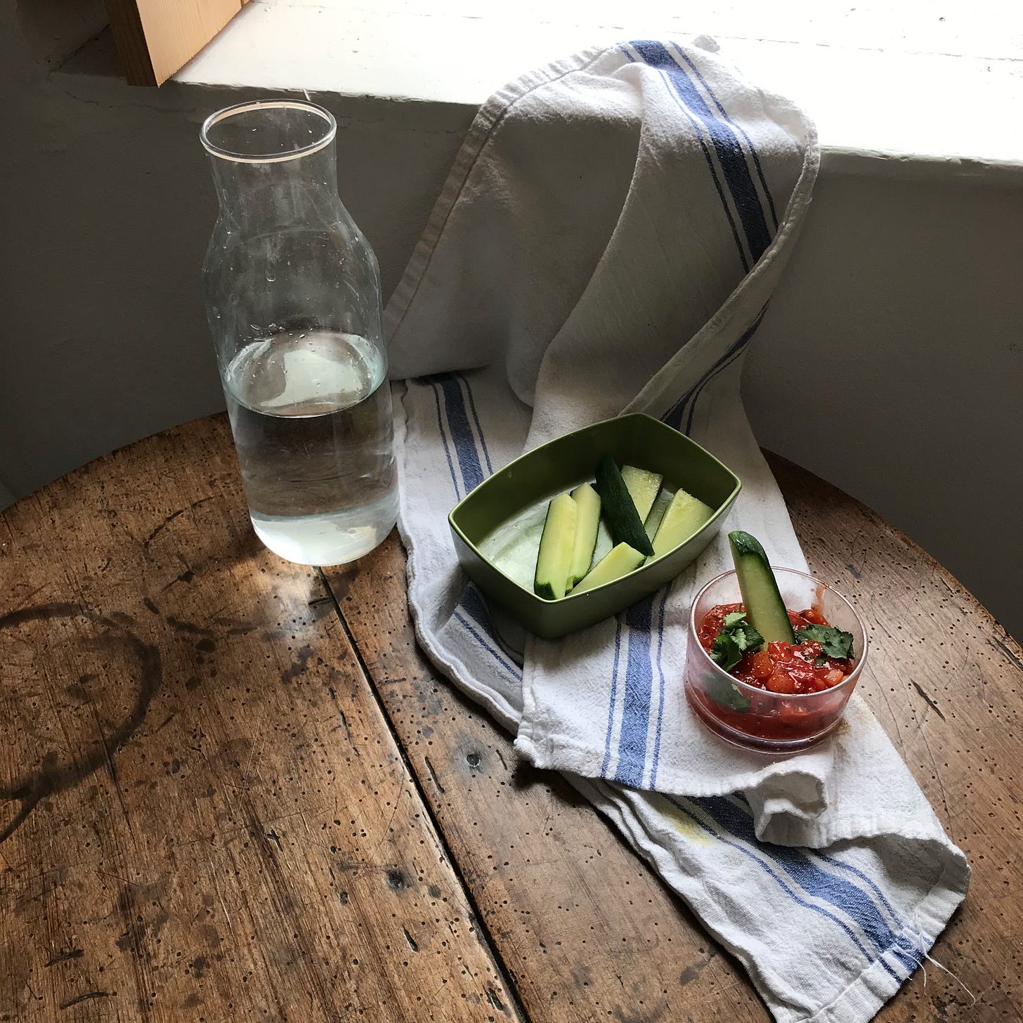 A red salsa in one round dish, and green batons of cucumber, on the same table, now bare and dark brown, with water, with a tea towel draped.