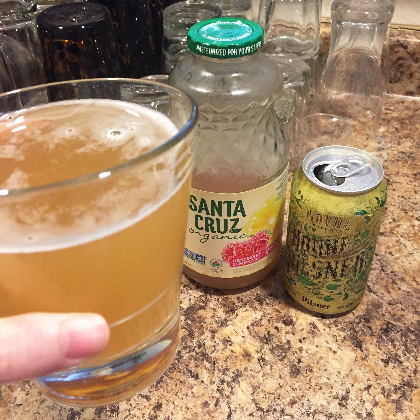 in the foreground, my hand holds a lightly foamy glass of shandy. A bottle of Santa Cruz raspberry lemonade and an open can of Hoyne pilsner are visible in the background.