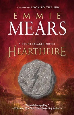 Cover of the book "Hearthfire" by Emmie Mears. The cover features the title, author, the phrases "Author of Look To The Sun" and "A Stonebreaker Novel," and the blurb "Masterful storytelling." by Ellie Ann, New York Times Bestselling Author. The cover art is an illustration of a small gray stone with a lightning bolt carved into it, pictured against a blurry wetland landscape with a red-tinged sky.