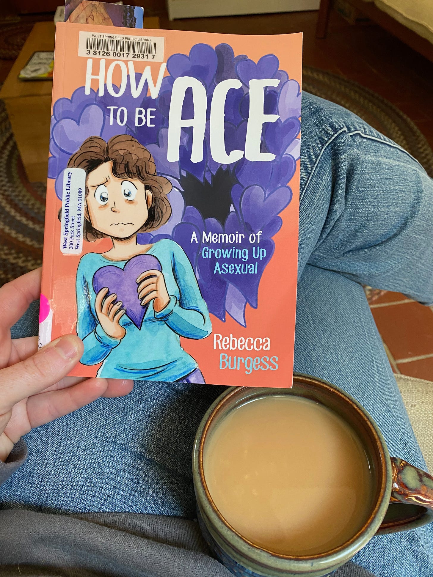 My hand holding a paperback copy of How to be Ace by Rebecca Burgess. My legs are crossed and there is a mug of tea in my lap.