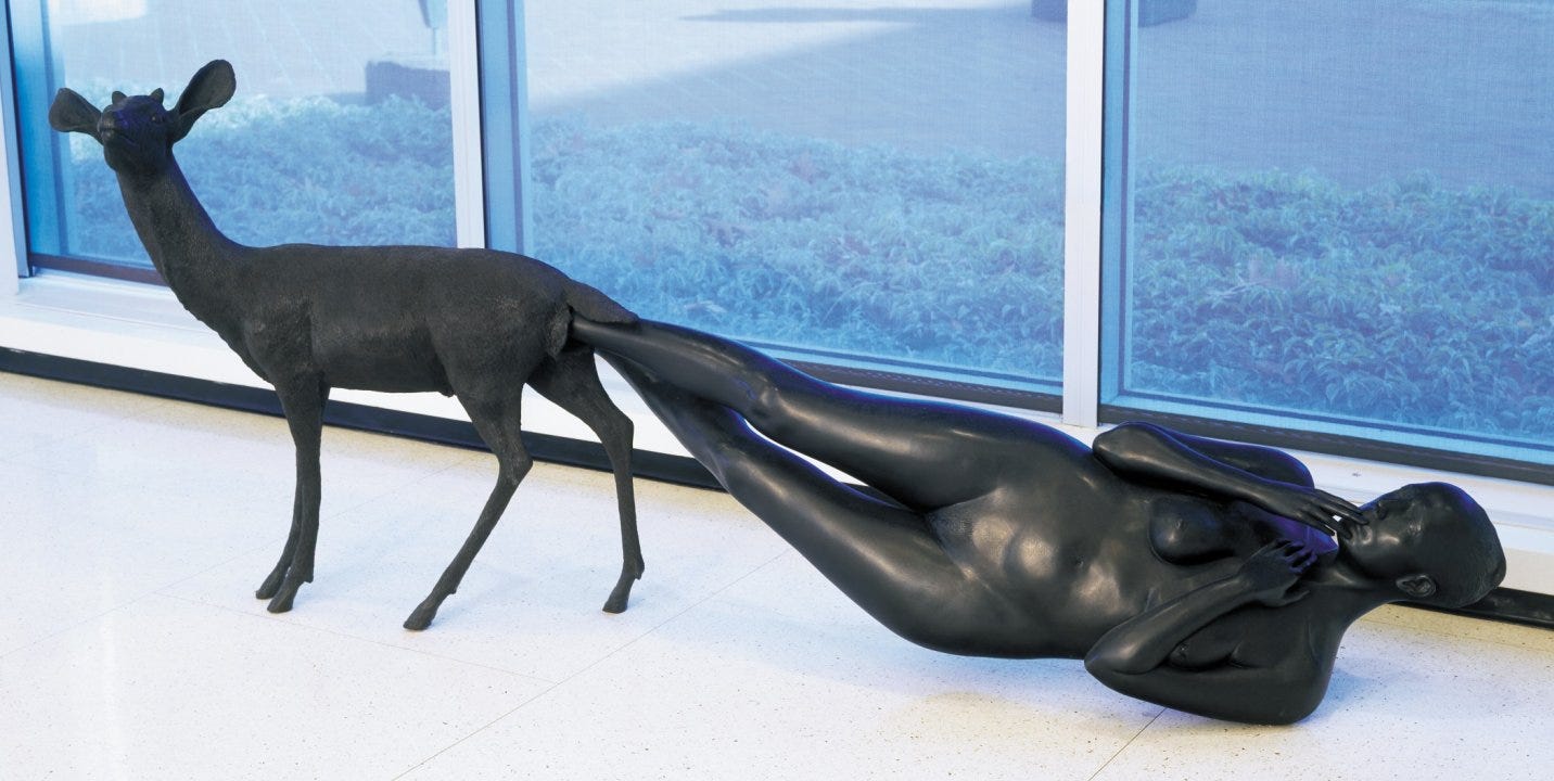 A bronze sculpture of a metal deer giving birth to an adult biological woman. The deer is on the left of the image, and the women she is birthing lies on the floor to the right - feet still within the deer, and her hands reaching towards her face. The sculpture is situated near a several windows, on a white tiled floor.