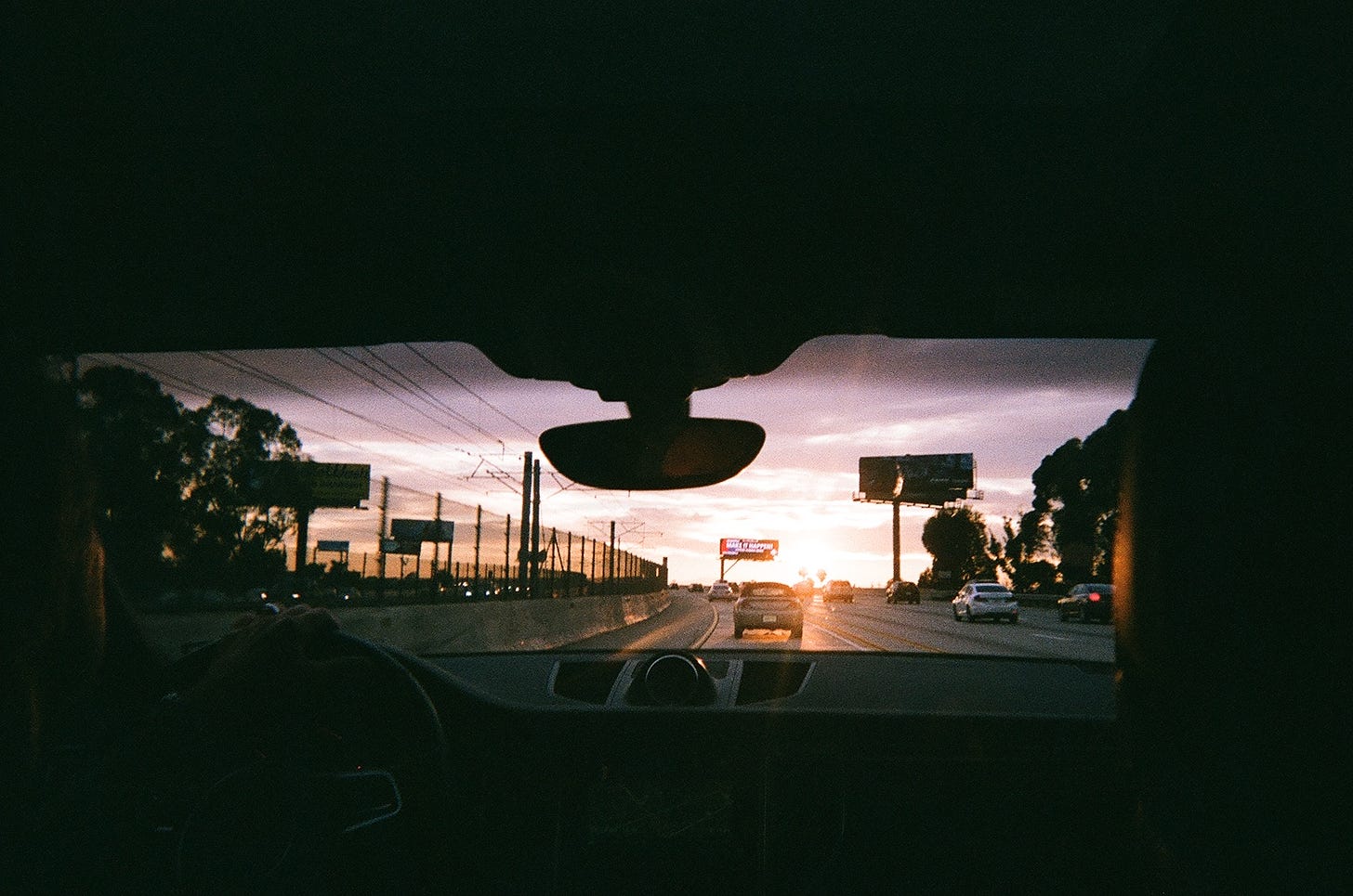 A Los Angeles freeway at sunset through the front windshield of a car, as seen from the car's backseat.