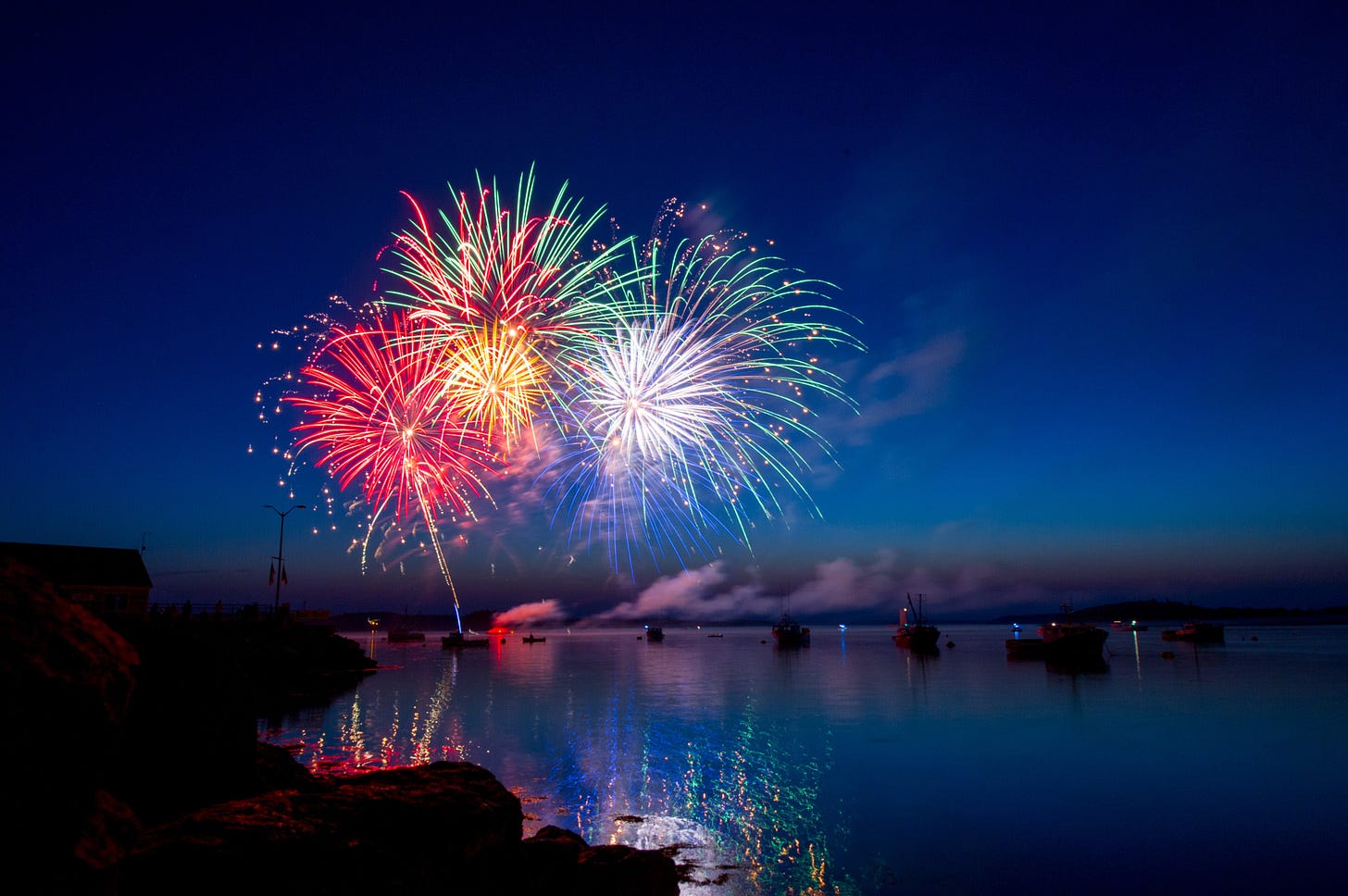 Multi coloured fireworks exploding against a dark blue sky over calm dark blue water.