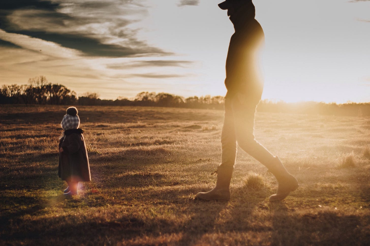 A father walks behind a small child in a field as the sun sets.