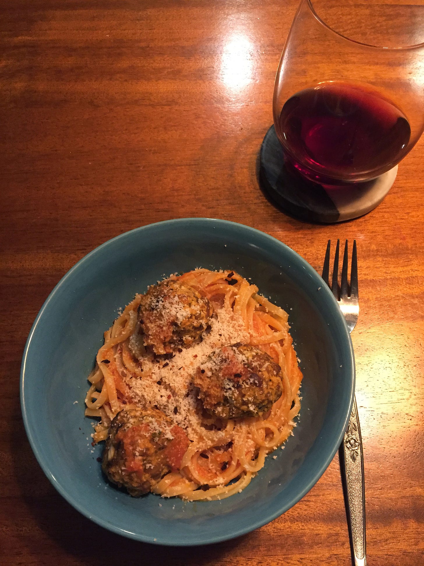 A blue bowl of linguine in tomato sauce with three tempeh meatballs on top, dusted with parmesan and chili flakes. A glass of red wine is on a stone coaster in the upper right.