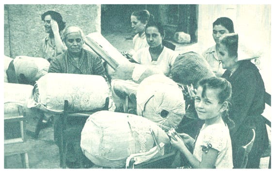 A group of women and girls around big, cylindrical cushions used to make lace. It's a black and white photo from the 1950s.