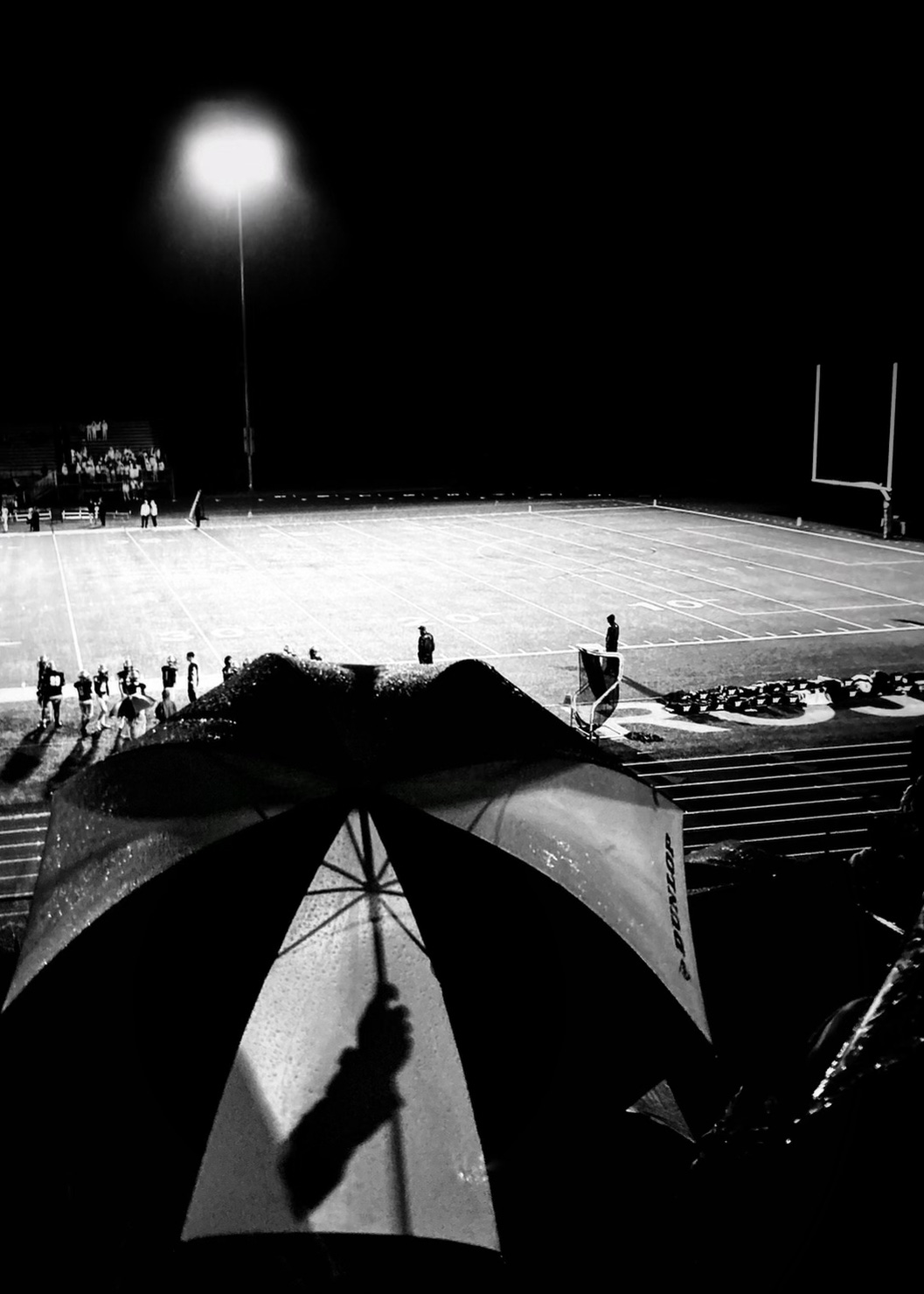 A woman shield by an umbrella rises to her feet in the stands of the football game. A silhouette of her arm holding the umbrella is seen through the backlit umbrella.