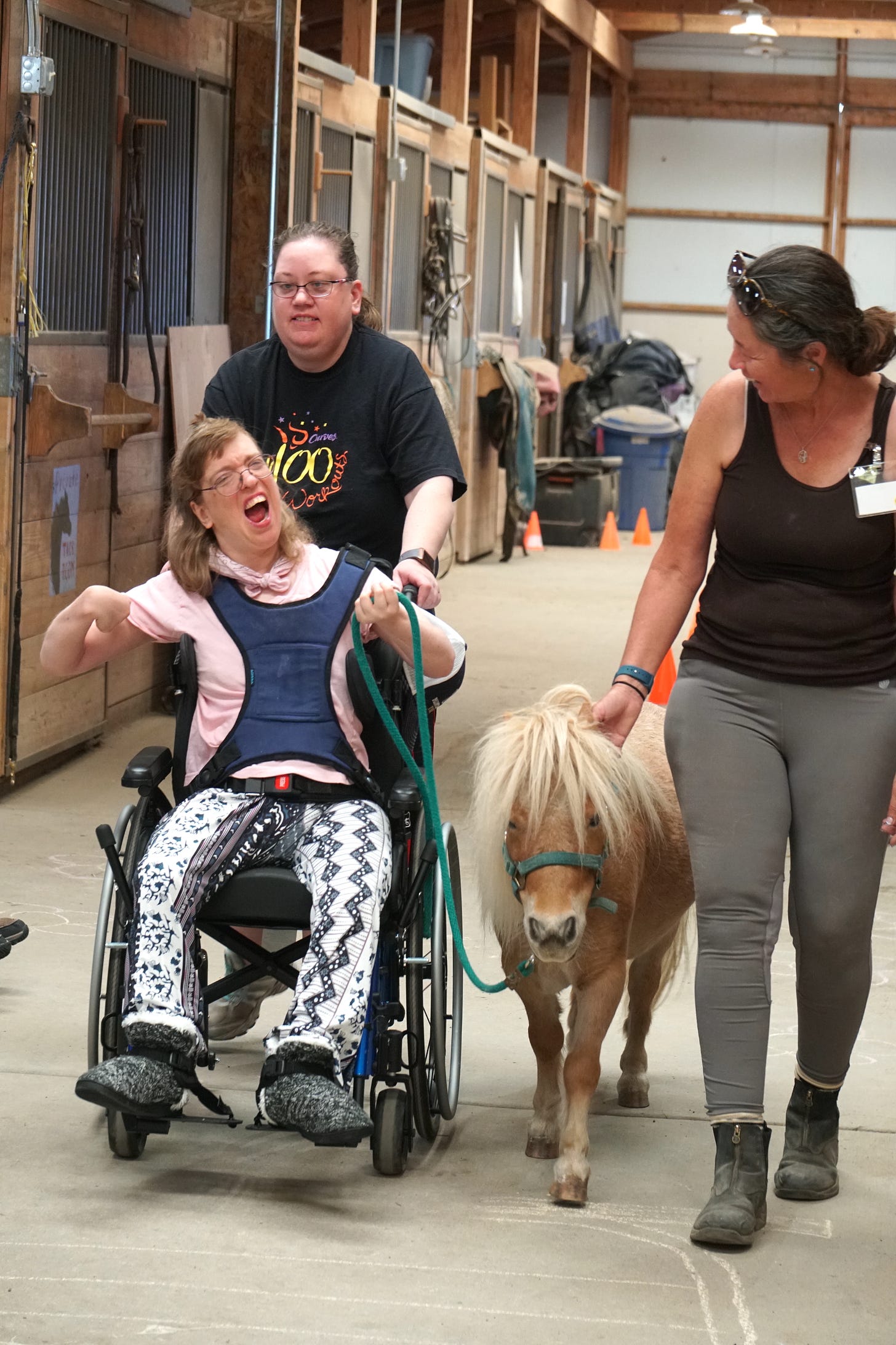 A woman in a wheelchair wearing glasses and a harness to keep her upright smiles and laughs as she leads a miniature horse through the Eagle Mount stables.