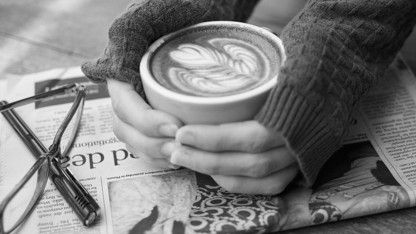 Two hands hold a coffee cup with latte art resting on a newspaper. Via Pxfuel