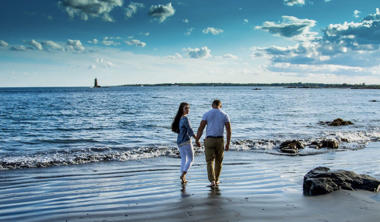 A couple walks along a beach on a sunny day. A lighthouse stands in the distance.