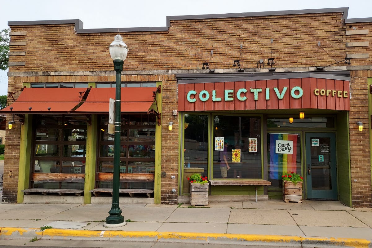 A front view of an aged one-story brick building. A rust colored wooden overhang over a green door next to sage green framed display windows has a sign for the Colectivo Coffee company. The letters are block. To the left in the forefront is a vintage green poled street lamp. Behind that is a second display window in the form of rolling garage doors under a rust colored canopy. Benches have been built into the door openings.