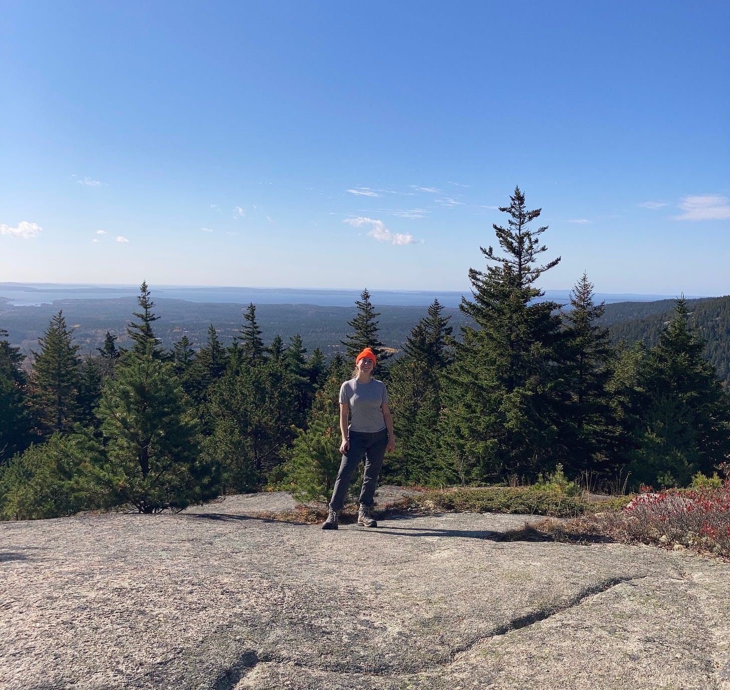 Kate, wearing a blaze orange knit cap and hiking clothes, stands on a rocky mountain smiling at the camera. Behind Kate are evergreens, and behind the evergreens is a view to the horizon of islands and water.