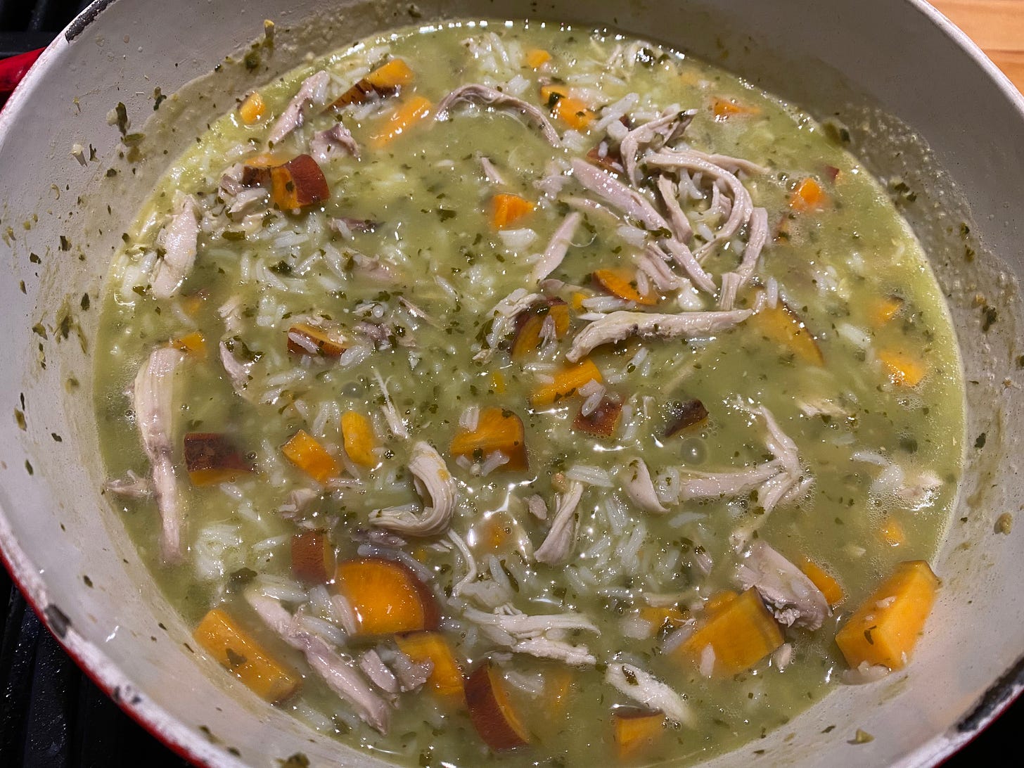 Close up of a pot of soup on a stove. The broth is green, with flecks of cilantro, lots of shredded chicken, and small cubes of sweet potato.