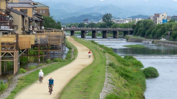 Bike riders along the Kamo River in Kyoto, mountain backdrop, old houses on stilts