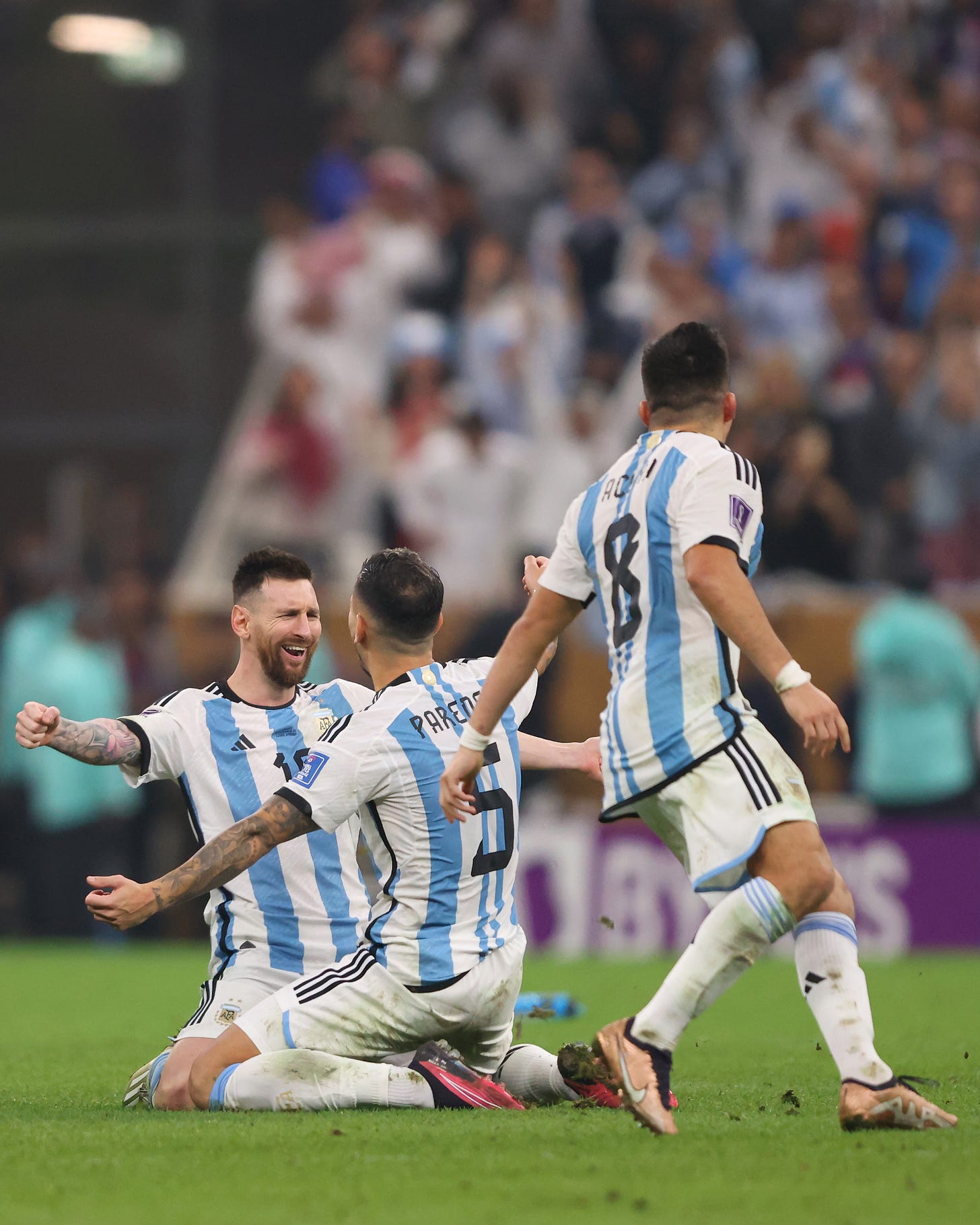 Leo Messi ajoelhado, sendo abraçado por Paredes; ao fundo, imagem desfocada da torcida. Foto logo após o pênalti que deu a Copa do Mundo de 2022 à Argentina.