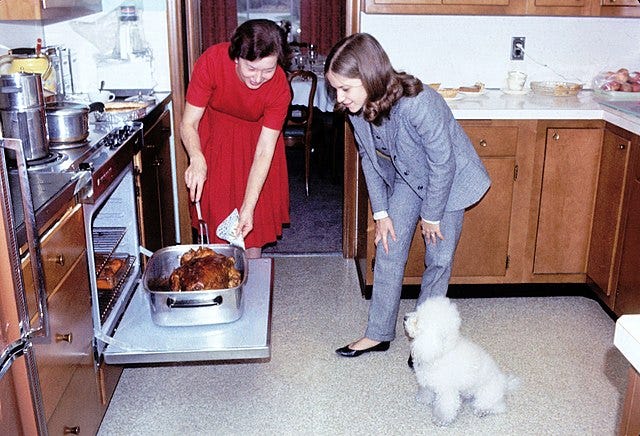 A woman in a red dress bends down to stick a fork into a turkey while a young woman wearing a suit looks on. The photo is from the 1960s. 