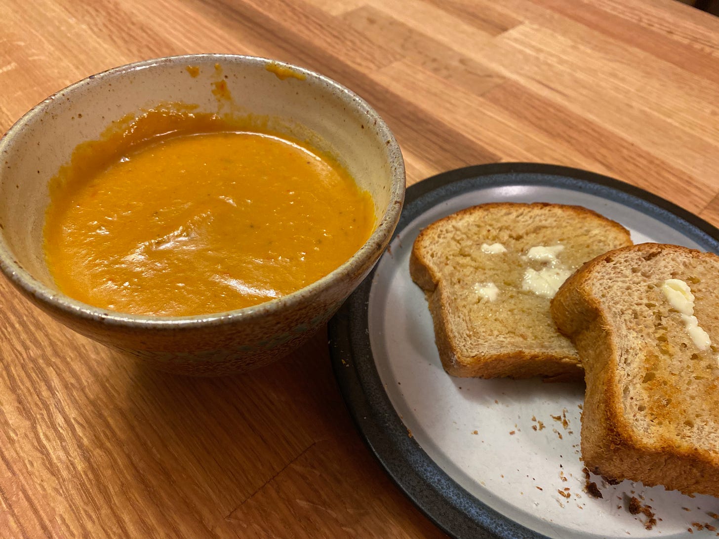 A ceramic bowl of purred orange soup sits on a wooden counter next to a plate holding two slices of buttered toast.