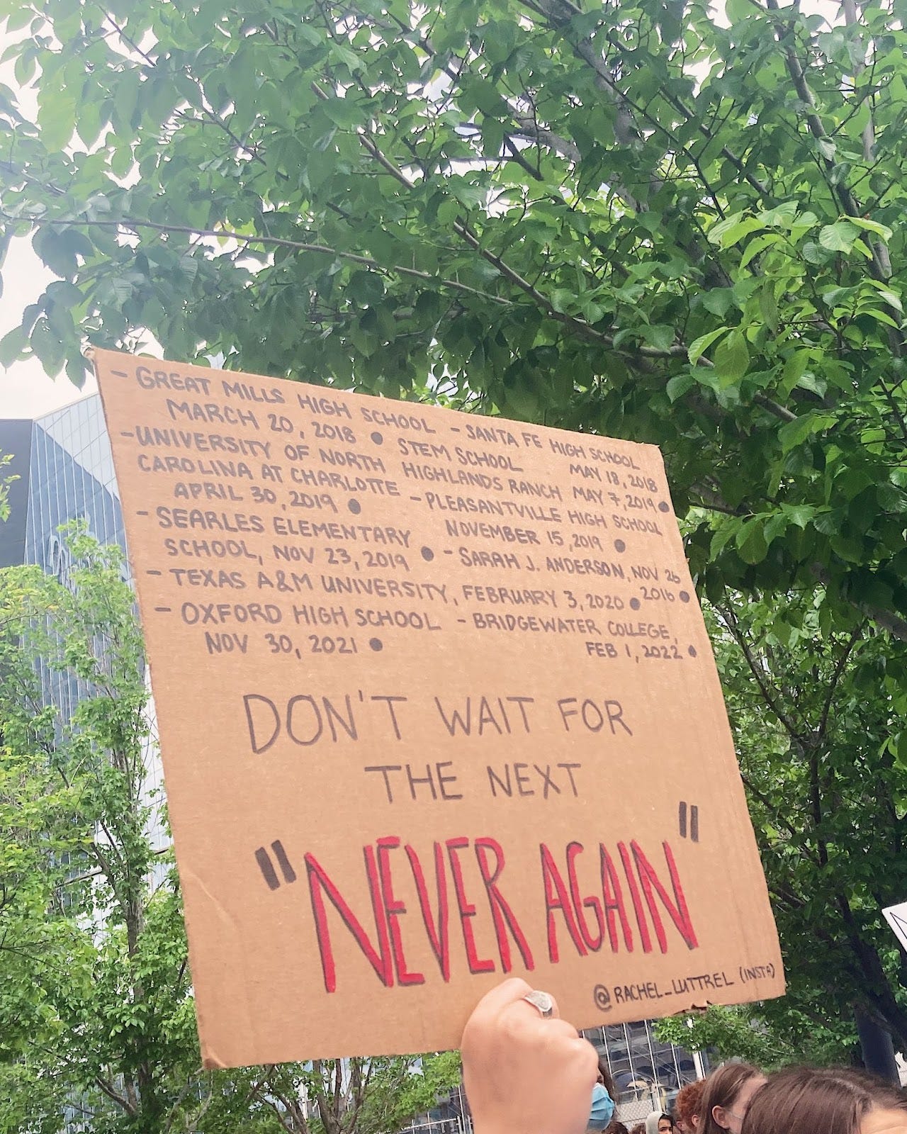a hand holds up a cardboard sign displaying the names of school shootings and "don't wait for the next" "never again"