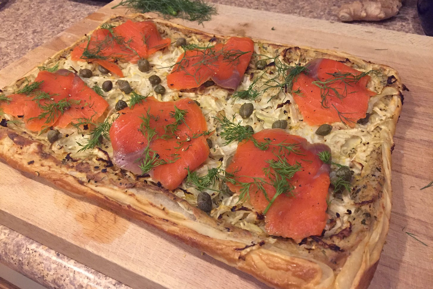 on a wooden cutting board, a puff pastry tart with fennel and cheeze on the bottom, topped with slices of smoked salmon, capers, and springs of fresh dill.