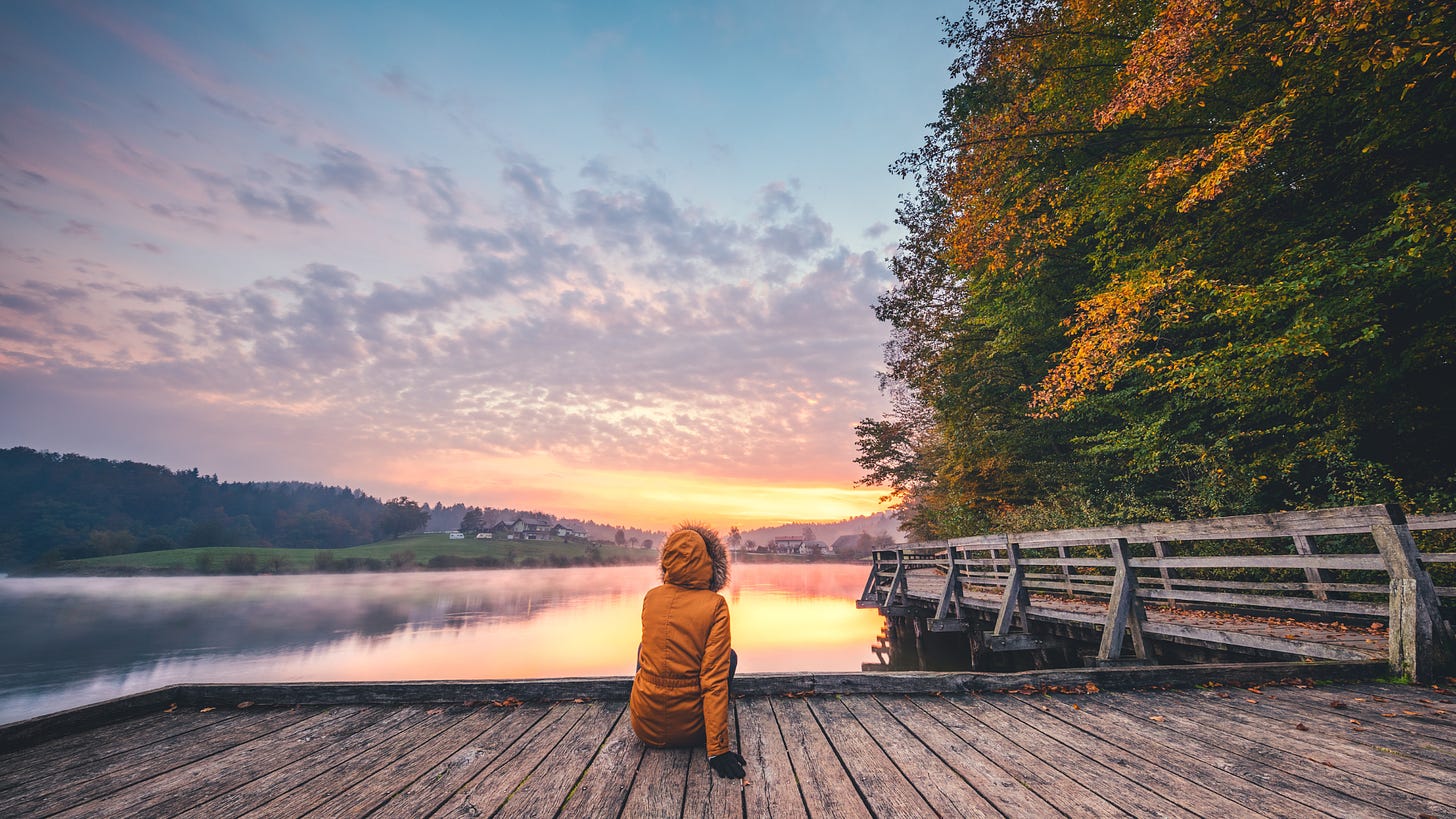 Person in orange winter jacket sitting on wooden deck overlooking a lake and colorful sunset