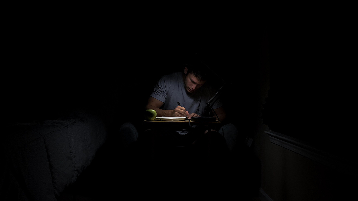 Man sitting in the dark, writing, for article titled “The Book That Never Gets Written” on The Reflectionist