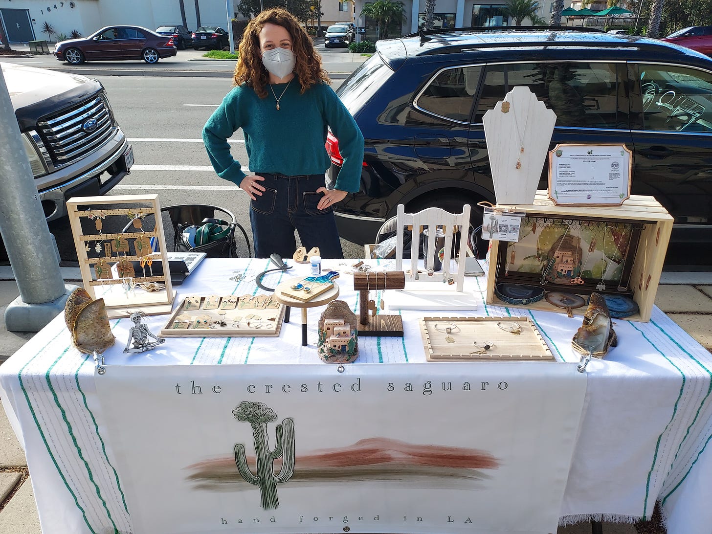 A woman stands behind her table at a local makers market. The table is covered in a white table cloth and various jewelry and metal pieces.