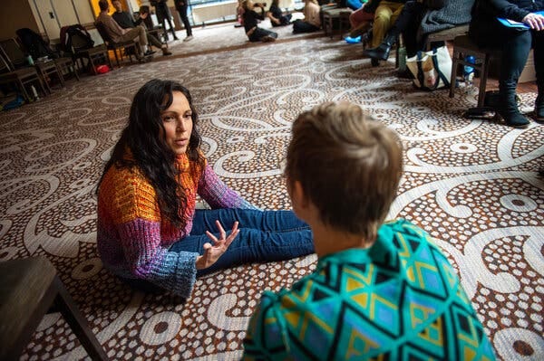 A woman in a knit orange, purple and blue sweater sits on a patterned rug in a hotel conference room, speaking to another person seated on the floor. In the background, several other program participants sit in chairs lined around the room.