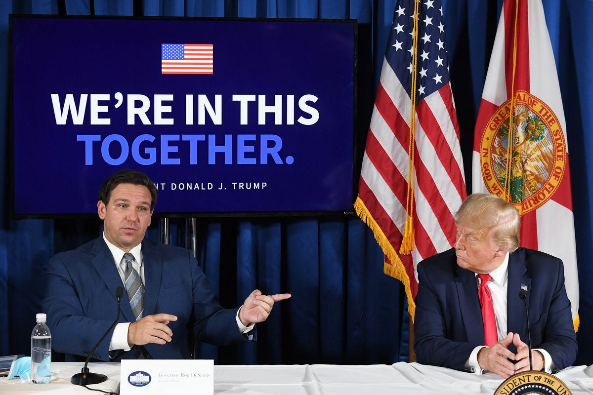 Governor DeSantis sits at a table pointing to his left at Donald Trump. Behind them in an American flag and a sign reading “We’re in this together.”