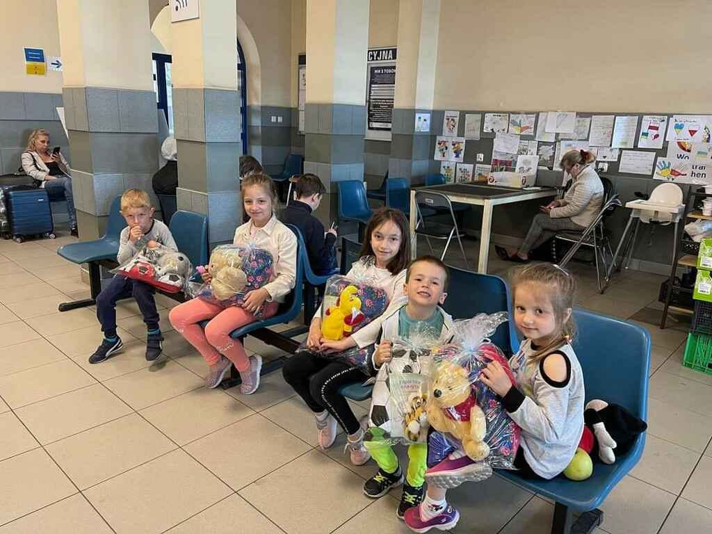 Five children smile at the camera from their seats in the Lublin Train Station. Each has a bag on their lap filled with a stuffed toy and a blanket.