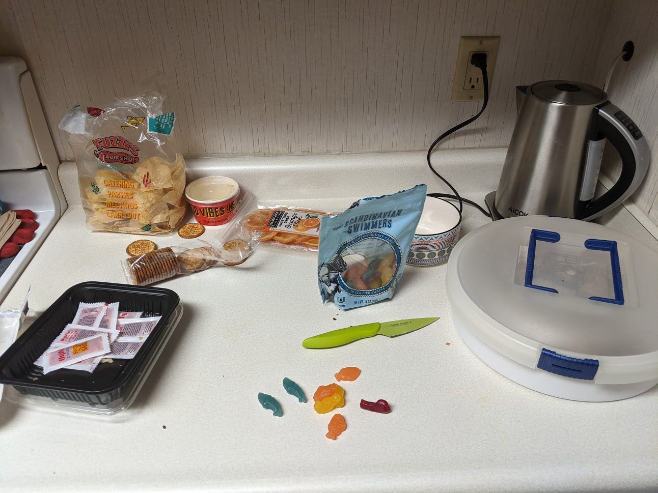 Various left-over snack items look a little bit squalid on the countertop; there is a small, green paring knife looking vaguely ominous next to several scattered Trader Joe's brand Scandinavian Swimmers.