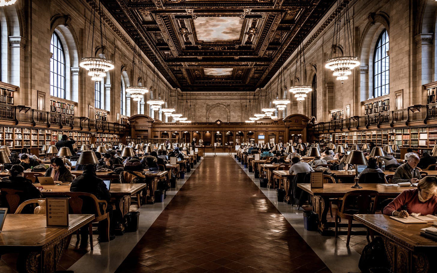 The Rose Main Reading Room at the Stephen A. Schwarzman Building of the New York Public Library, filled with people reading at wooden tables, under chandeliers. 