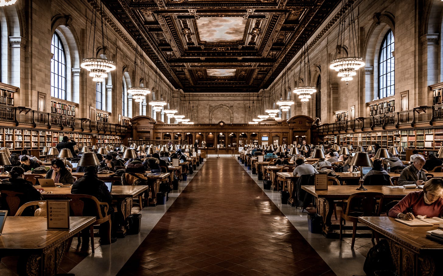 The Rose Main Reading Room at the Stephen A. Schwarzman Building of the New York Public Library, filled with people reading at wooden tables, under chandeliers. 