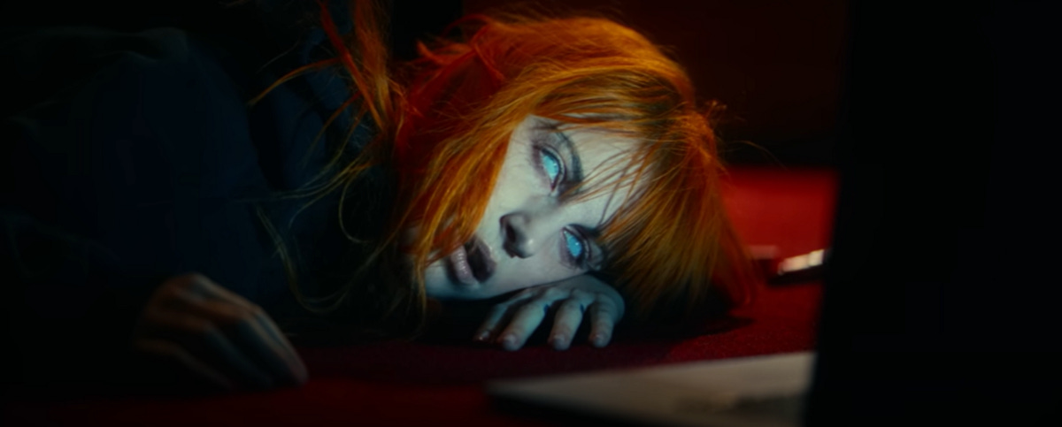 A woman with orange hair and bangs — Hayley Wiliams — lies on the floor in front of a laptop screen. Her eyes are glazed over, appearing white.