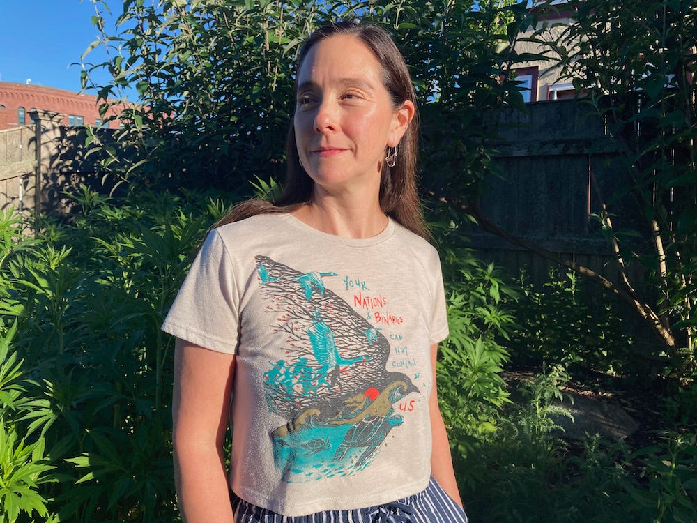 A mixed-race person with long brown hair stands in front of a stand of green-leafed plants. Her gray croped tee shirt features a design of flying birds, swimming turtles, and running people.