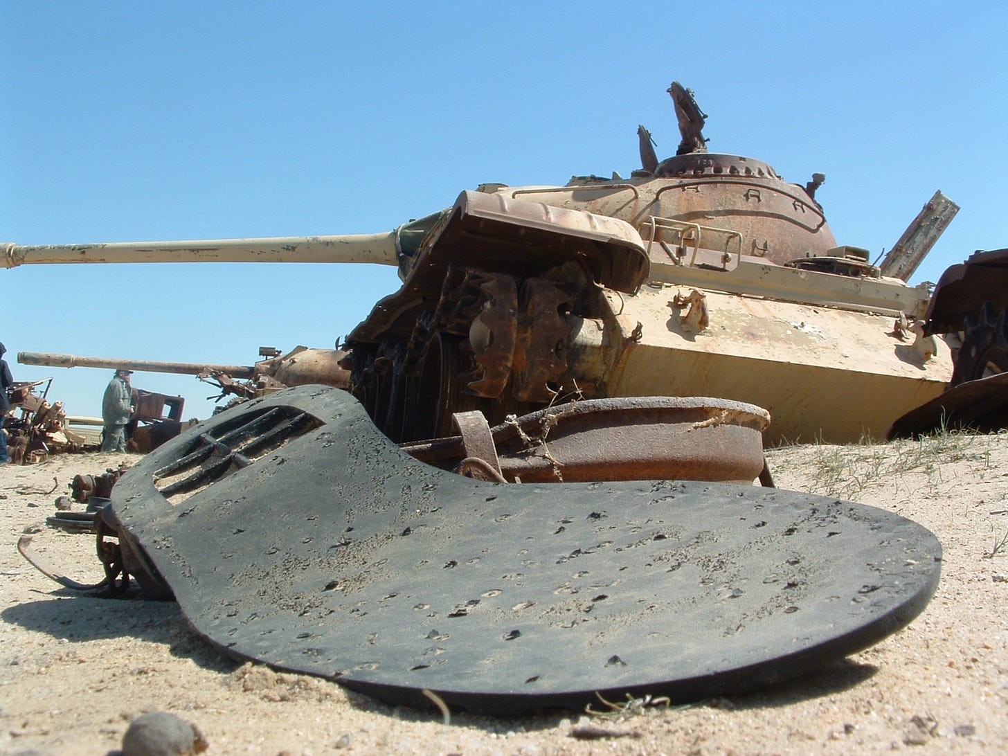 The sole of a shoe can be seen in the foreground. Behind it are the wrecks of multiple tanks, and other ruined military machines, blasted and weathered.