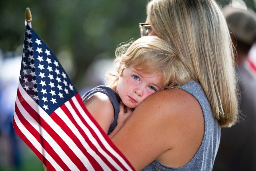 A woman holds a small child in her arms, standing among others, beside an American flag.