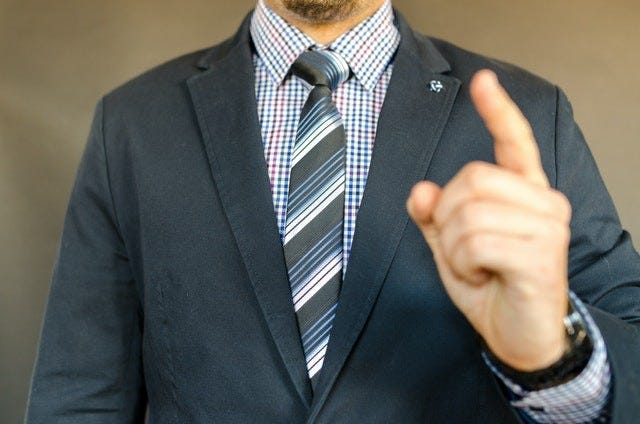 close up of a person in a suit from the chin down. the person is raising a pointed finger as if giving a demand or admonition