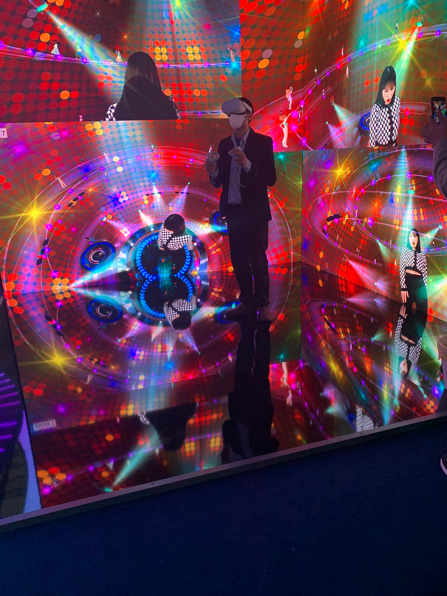 A man in a suit wears a mask, VR headset, and hand controllers while he stands in a technicolor cube (missing three sides, which is how we can see into it).