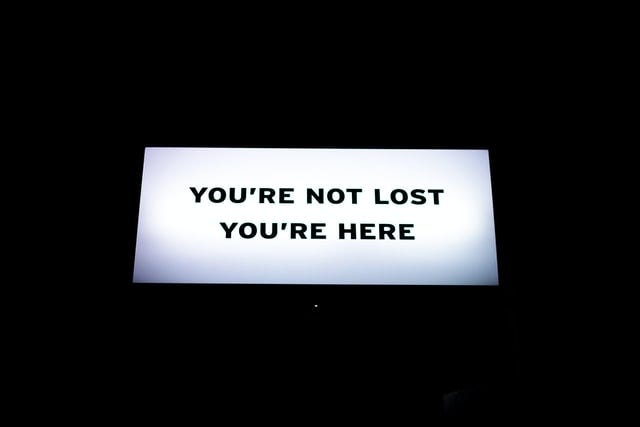 Photo. White neon sign with black text: "You're not lost, you're here."