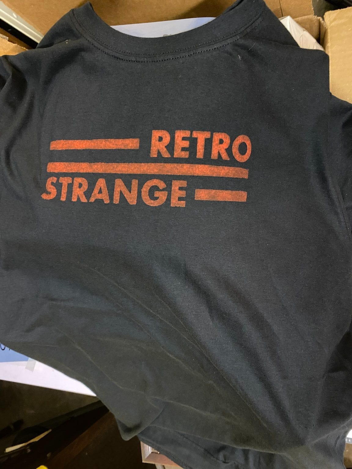 A photograph of a black t-shirt with the RetroStrange logo printed on it in orange.
