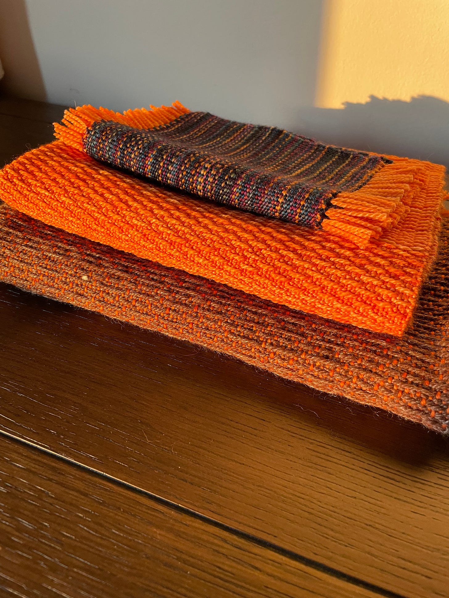 Three handwoven pieces folded and sitting in golden hour light. The top is multishaded blue, the second is all orange with long floats, and the bottom is brown with the orange warp showing through.