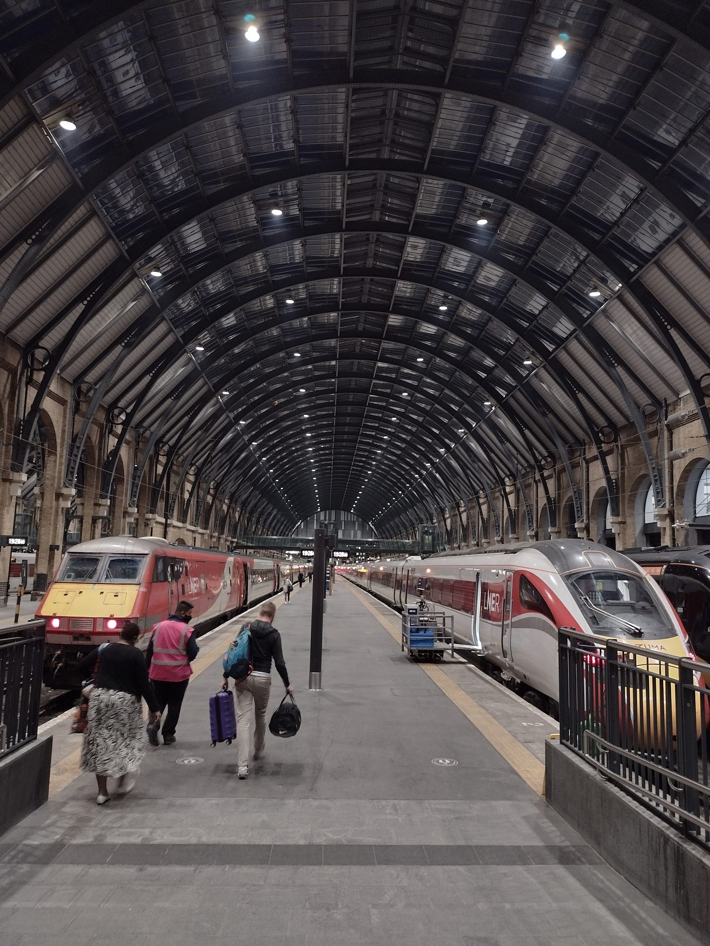 A photo of Kings Cross station. Two passengers are walking down the ramp. Two trains wait either side of a central platform. The roof arches overhead