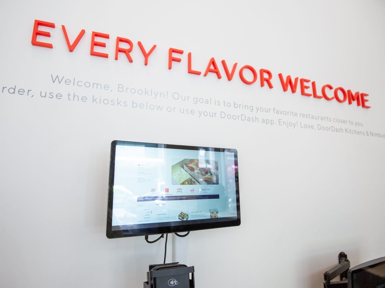 A wall-mounted touch screen with a card payment pad underneath. Red text on the wall reads "every flavor welcome."