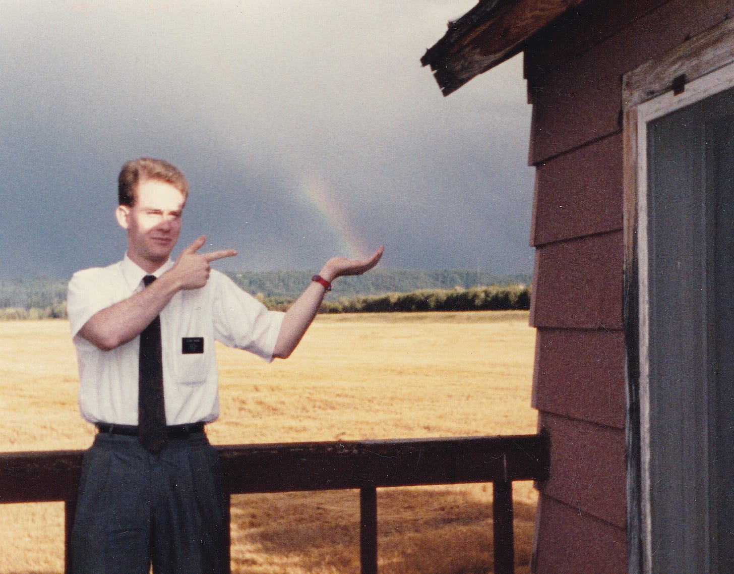 A Mormon missionary poses on the deck of a tar-shingled house with a wide wheatfield behind him. It looks like he's catching a distant rainbow in his open hand.