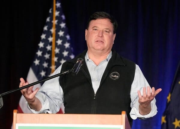 Todd Rokita wearing a gray collared shirt and a black vest. He gestures with his hands behind a lectern.