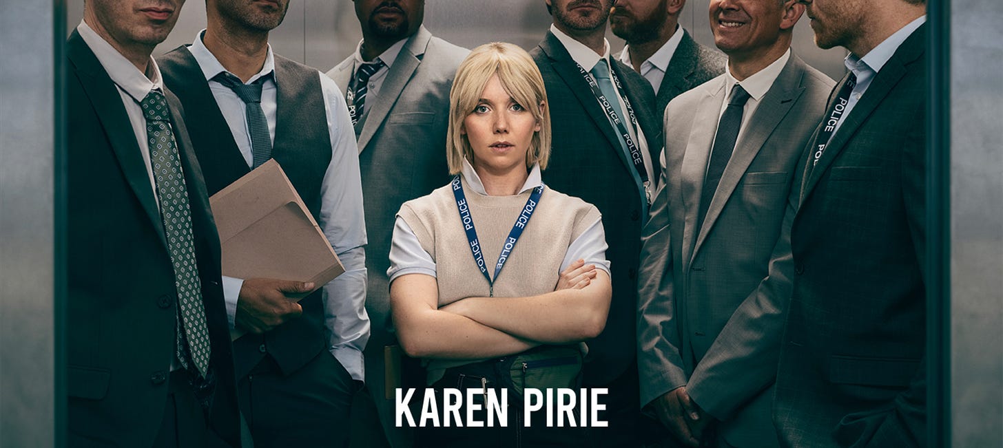 A blonde white woman wearing a tan sleeveless top with a collared white short-sleeved blouse underneath stands with her arms crossed in an elevator surrounded by many men in suits and a name reading "Karen Pirie" imposed on top.