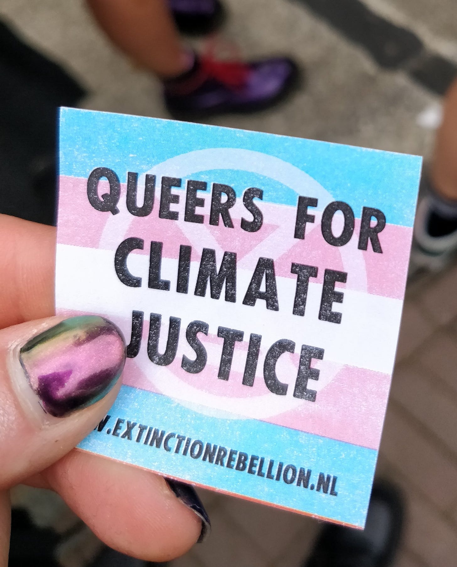 A hand holds a sticker that says 'Queers for Climate Justice' with a link to www.extinctionrebellion.nl at the bottom