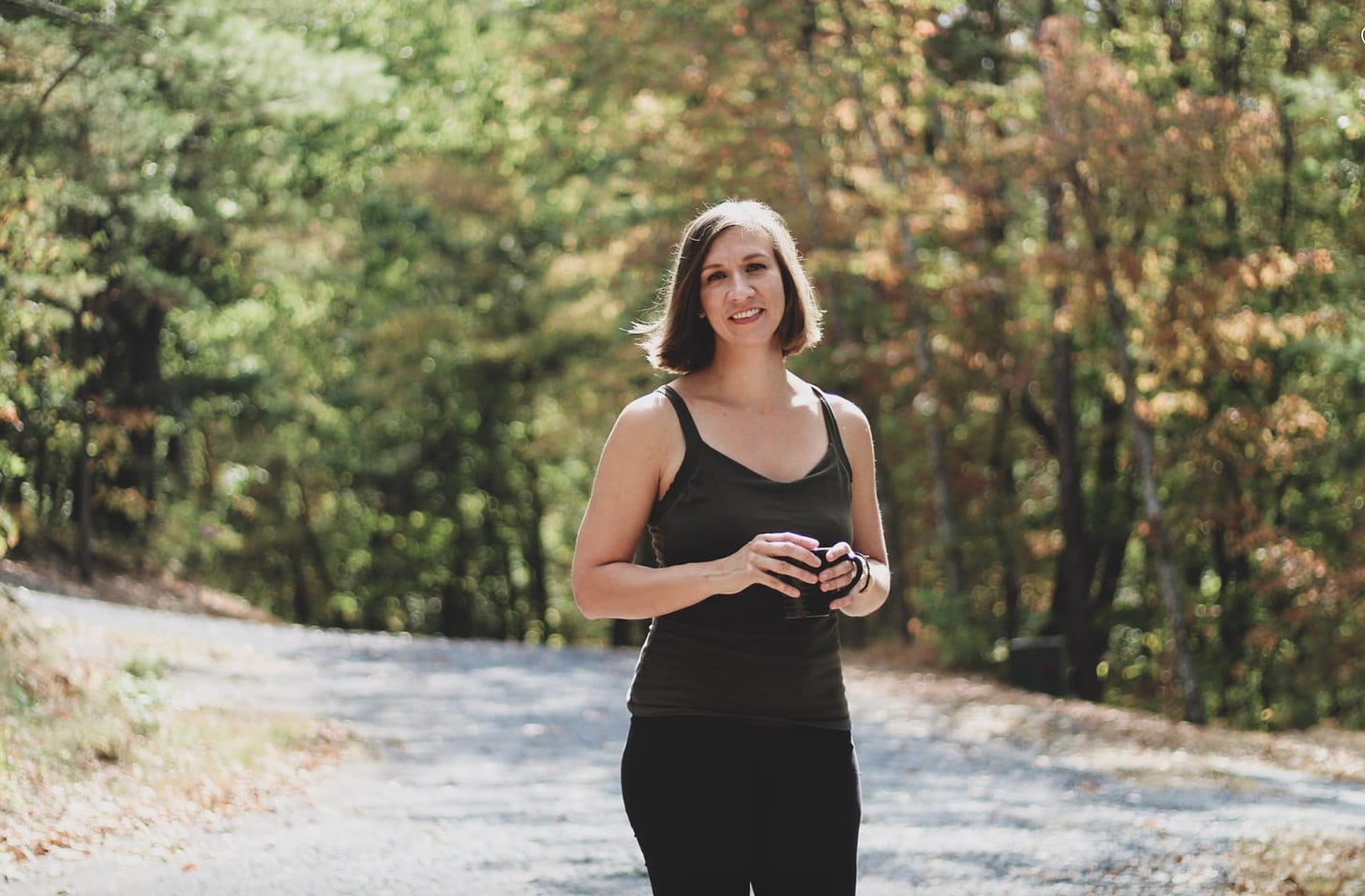 A white, brunette woman in an olive tank top and black pants holds a black coffee mug on a trail in the woods