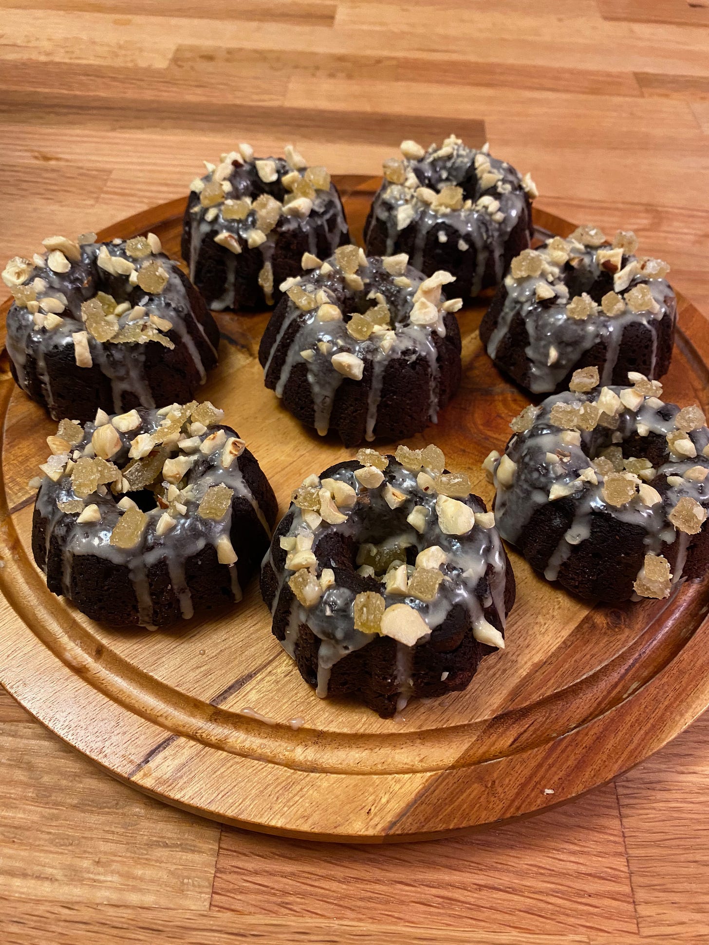 Eight mini chocolate bundt cakes are arranged in a circle on a wooden platter. They all have vanilla glaze running down their sides, and they are topped with chopped candied ginger and hazelnuts.