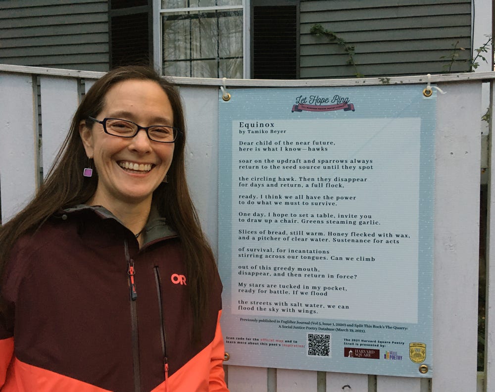 Me (mixed race woman with long brown hair and glasses) Standing next to a plastic plaque with my poem "Equinox" hanging on a white fence.